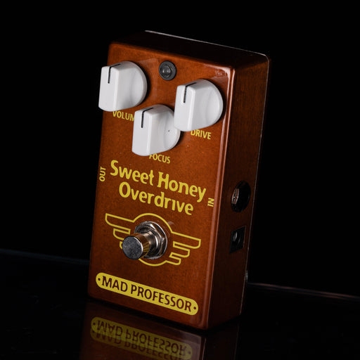 A Sweet Honey Overdrive guitar pedal by Mad Professor, featuring a brown wood-grain finish, three white rotary knobs, and a silver footswitch on a black reflective background.
