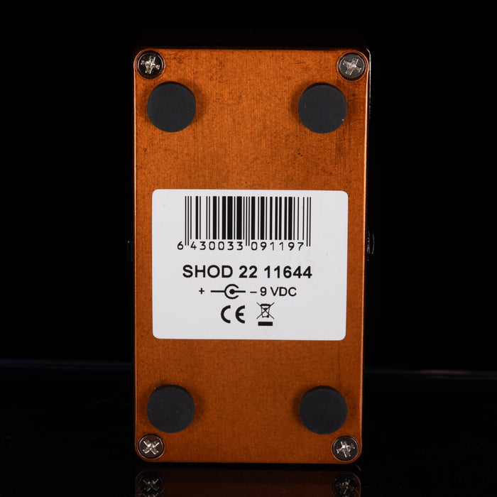 The back of a copper-colored guitar effects pedal with four black rubber feet and a white barcode label in the center, shown against a black background.