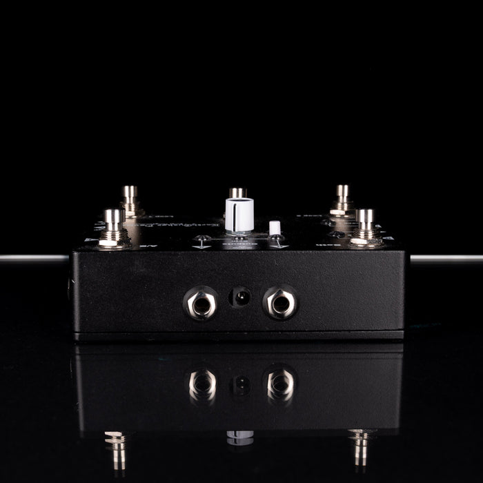 Black electronic pedal with various knobs and ports on a reflective surface with a black background