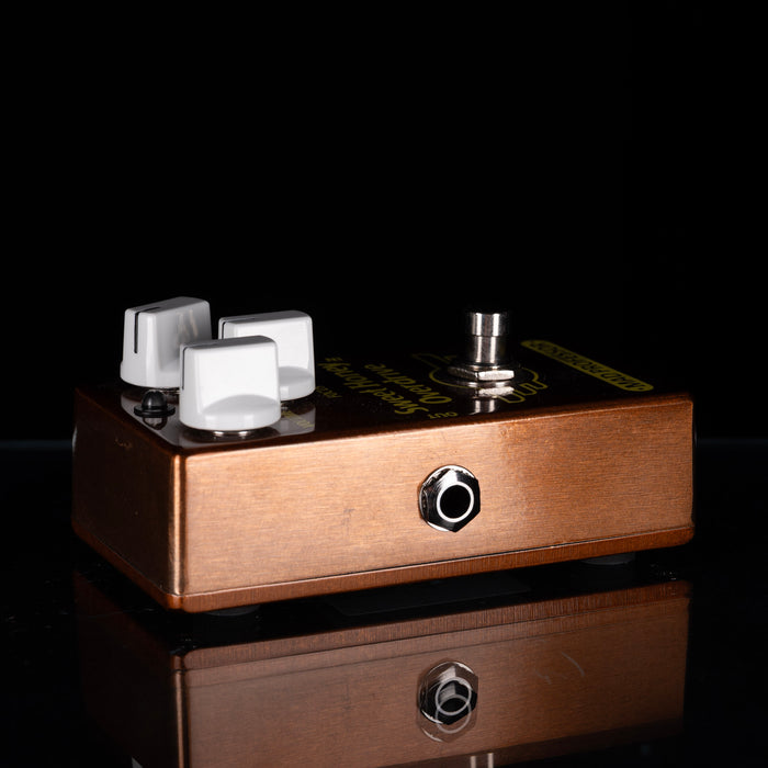 A copper-colored guitar effects pedal with three white control knobs, a silver footswitch, and gold lettering, shown on a reflective black surface against a black background.